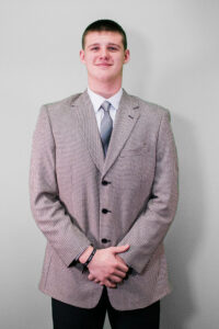 a man in a suit and tie stands with his hands folded smiling at the camera