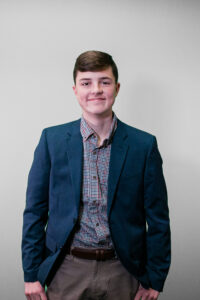 a young man wearing a blue jacket and a plaid shirt standing with his hands in his pockets