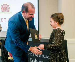 a man in a suit shakes hands with a little girl in front of a charter institute sign
