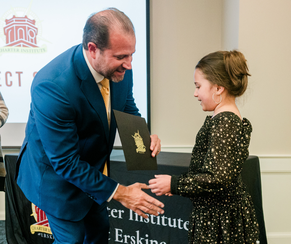 a man in a suit shakes hands with a little girl in front of a charter institute sign